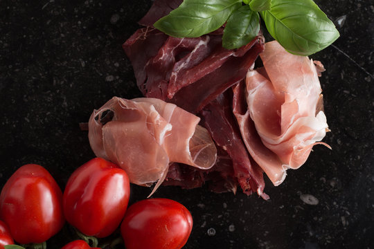 Jambon mix. Ham. Traditional Italian and Spanish salting, smoking, dry-cured dish - jamon Serrano and prosciutto crudo sliced with herbs and tomatos on dark stone background. Copy space. Closeup. 