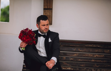 Stylish groom sitting on a bench. Groom in black suit with boquet of red piones in hand. Free space.