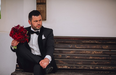 Stylish groom sitting on a bench. Groom in black suit with boquet of red piones in hand. Free space.
