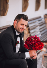 Stylish groom sitting on a bench. Groom in black suit with boquet of red piones in hand. Free space.