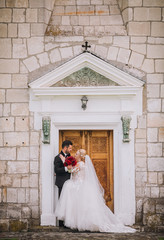 Romantic groom hugging beautiful bride near old church with big wooden doors.