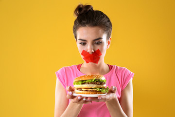 Woman with tied mouth holding hamburger on yellow background
