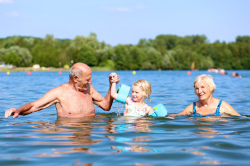 Happy kid swimming in the lake. Healthy smiling toddler girl learning to swim wearing inflatable armbands. Granddaughter enjoying summer vacation with grandparents.