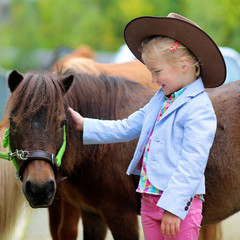 Lovely cowgirl caressing little pony horse in the farm. Pretty preschooler girl wearing cowboy hat...