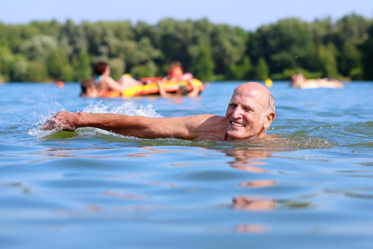 Healthy Senior Man Swimming In The Lake Or River. Happy Elderly Man Enjoying Active Summer Vacation. Sportive Lifestyle. Active Retirement Concept.