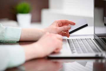 Business woman in a green blouse sitting at the desk in the office and working on laptop computer with a credit card in her hands .