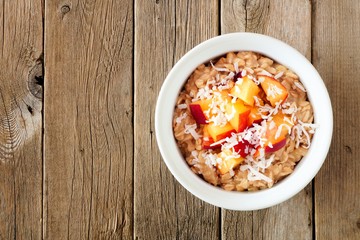 Bowl of overnight breakfast oats with diced peach and coconut, overhead view on rustic wood