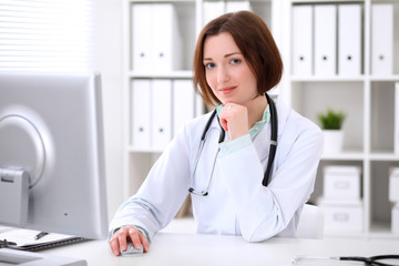 Young brunette female doctor sitting at a desk and working on the computer at the hospital office.  Health care, insurance and help concept. Physician ready to examine patient