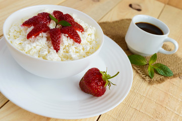 Homemade cottage cheese with fresh strawberries, mint leaves decoration in a white bowl and a cup of coffee. Useful eco breakfast. Healthy eating.
