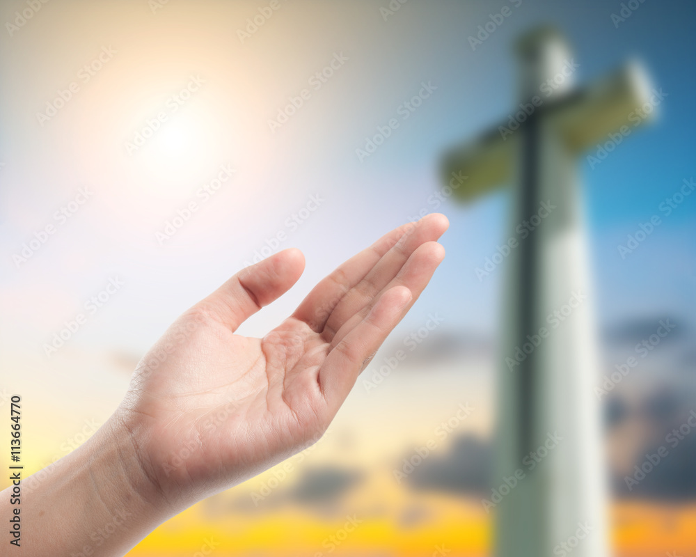 Wall mural hand praying over blurred the cross on the sky background.