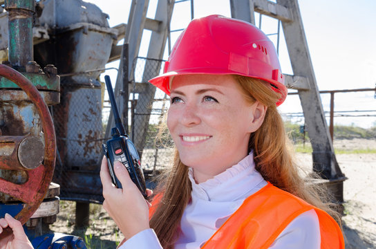 Beautiful woman engineer near well head valve in the oil field talking on the radio wearing red helmet and work clothes. Pump jack background. Oil and gas concept. 