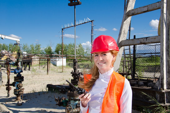 Beautiful woman engineer near well head in the oil field talking on the radio wearing red helmet and work clothes. Pump jack and pipeline background. Oil and gas concept. 