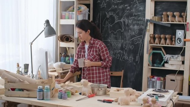 Locked-down shot of young female Asian artist drinking tea, using computer and putting on work apron at her handmade studio dealing with production of paper mache toys