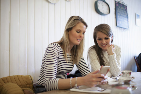 Two Young Female Friends Check Mobile Phones In Tea Shop