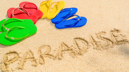 Fototapeta na wymiar Colored sandals fanned out on a deserted tropical beach with the word paradise written in the sand