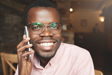 African American student wearing oval glasses, talking on mobile phone, looking at the camera with happy expression while sitting at a cafe against wooden wall background. Lifestyle and people concept