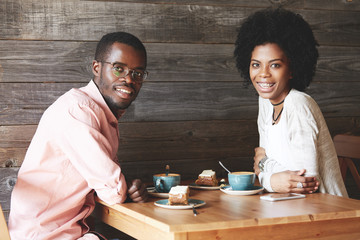 Young African office worker in glasses having lunch with his fashionable female colleague with Afro hairstyle and braces, looking and smiling at the camera while having a nice conversation at a cafe
