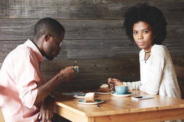 Beautiful young African woman with Afro hairstyle in stylish clothes looking at the camera while sitting at the table at a cafe with her boyfriend, having coffee and eating dessert during lunch