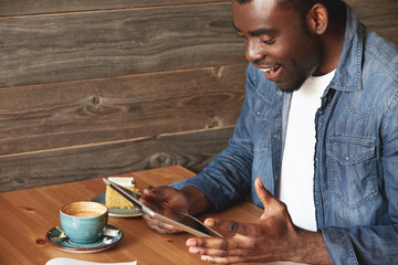 Rapturous African American man sitting in a caf?, enthusiastic about news or received messages from social networks. The guy is totally happy that forgot about his coffee and sweet pie lying on table.