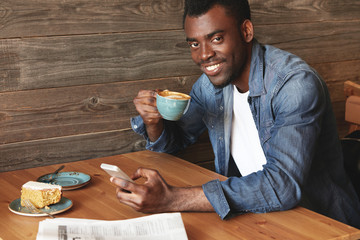 Handsome African American man checking feed on his device and drinking hot coffee. Young cheerful guy in blue denim shirt is enjoying his morning delicious cheese cake and recent news of the day.