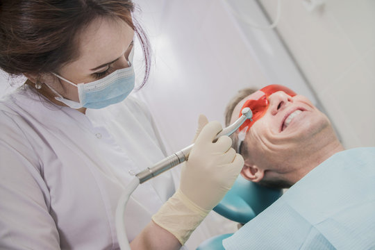 Dental Care Concept Stomatology Inspection. The Patient Lies In A Chair In Dentistry In Front Of Him A Hand With Drill. The Dentist Is In The Office Beside The Chair With The Patient