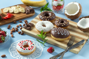 Delicious donuts. Served on the wooden board and white napkin. Decorated with strawberry, banana, coconut, mint and almond. Blue background. Horizontal image. With lemonade.