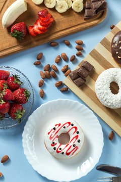 Delicious donuts. Served on the wooden board and white plate. Decorated with strawberry, banana, coconut and almond. Blue background. Silver knife and fork. Vertical image. Top view
