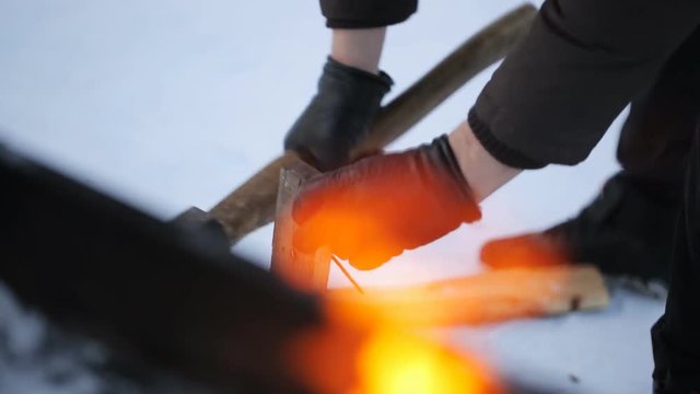 In winter, a man wearing gloves chopping wood with an ax on a background of fire.