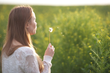 Young spring fashion woman blowing dandelion in spring garden. Springtime. Trendy girl at sunset in spring landscape background. Allergic to pollen of flowers. Spring allergy.