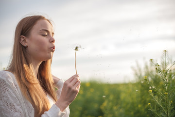 Young spring fashion woman blowing dandelion in spring garden. Springtime. Trendy girl at sunset in spring landscape background. Allergic to pollen of flowers. Spring allergy.