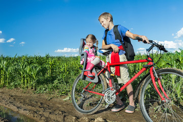 Fototapeta na wymiar Sibling teenage boy cycling with his little sibling sister on baby bike seat on farm corn field dirt road