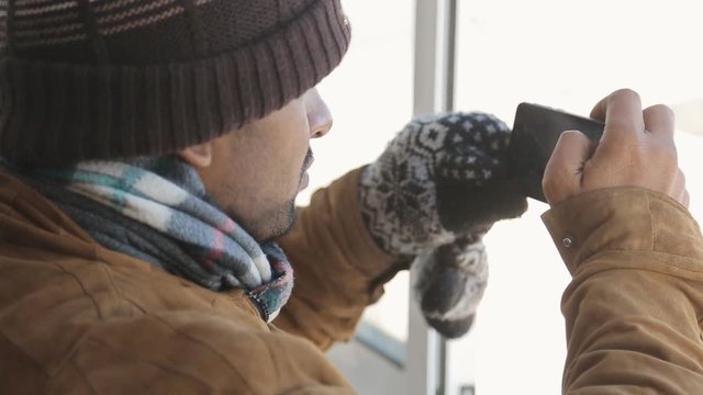 Serious warmly-dressed man-Hindu takes pictures with smart phone through a window in winter