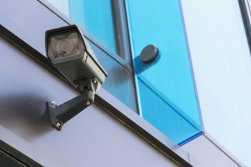 Grey security camera attached to wall blue windows in background