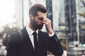 Frustrated young businessman looking stressed and tired while massaging nose on city street after work