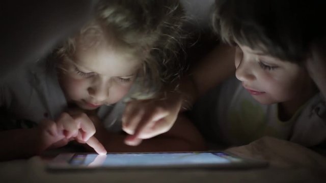 two little girls sisters draw on a tablet PC hiding under a blanket
