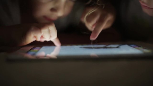 two little girls sisters draw on a tablet PC hiding under a blanket