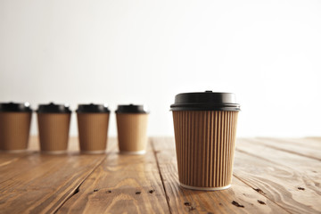 One focused cardboard paper coffee cup with black cap isolated on side in front of unfocused four take away cups on brushed vintage wooden table