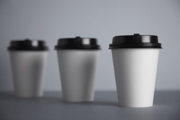 Three take away white paper cups with closed black caps, top view, isolated on simple gray background, first cup in close focus, cups behind are unfocused in bokeh