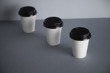 Three take away white paper cups with closed black caps, top view, isolated on simple gray background, first cup in close focus, cups behind are unfocused, top view