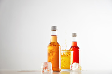 Obraz na płótnie Canvas Freezed in air splash of lemonade from felt ice cube into glass with yellow tasty drink between two sealed transparent unlabeled bottles with orange and red refreshment beverages isolated on white