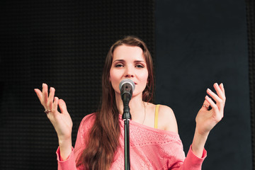 Fototapeta premium Young singing woman inro the microphone close-up. Beautiful woman in pink dress sings into the micriphone, hands are rised up. Songing woman looking at camera