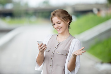 Portrait of smiling beautiful caucasian woman walking on the street and looking at smartphone screen with joyful expression. Casual female laughing at texts or video on phone outdoors in summer