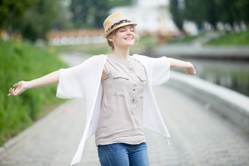 Portrait of casual traveler girl feeling ecstatic and alive on the street. Young joyful female breathing fresh air with closed eyes. Attractive model in straw hat relaxing with outstretched arms