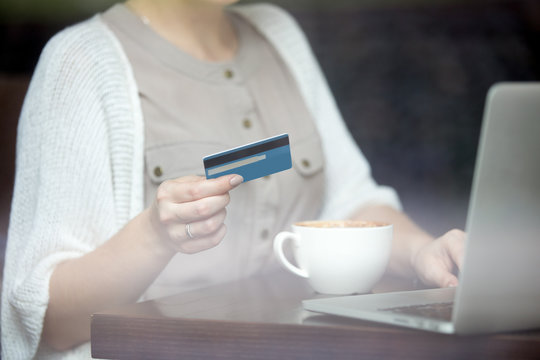 Young beautiful female sitting in modern urban cafe and paying with credit card for online shopping on laptop. Shot through window. Focus on plastic card. Close-up of hands