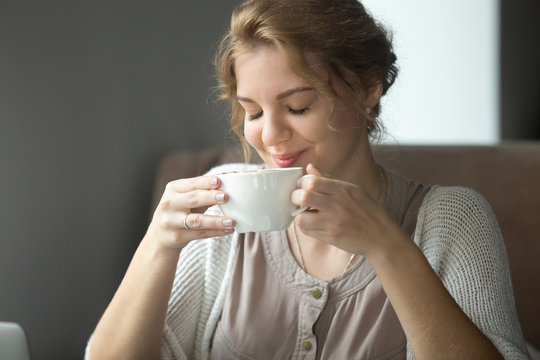 Half-length portrait of young beautiful female sitting at coffee shop with cup of tea or cappuccino. Woman enjoying hot drink in the morning. Attractive model sipping coffee with closed eyes