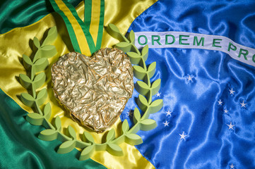 Large heart shaped gold medal surrounded by laurel wreath resting on shiny Brazil flag background with studio lighting