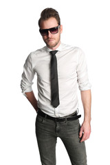A trendy focused man wearing dark sunglasses, a white shirt with a black tie and jeans, standing against a white background.
