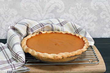Homemade pumpkin pie on a cooling rack with towel wrap.