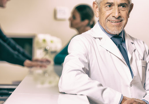 Smiling Senior Male Doctor Smiling At Hospital Reception Desk, F