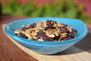 Blue glass bowl filled with trail mix with sliced almonds, salted sunflower kernels, and sugared raisins on a handmade wood cutting board